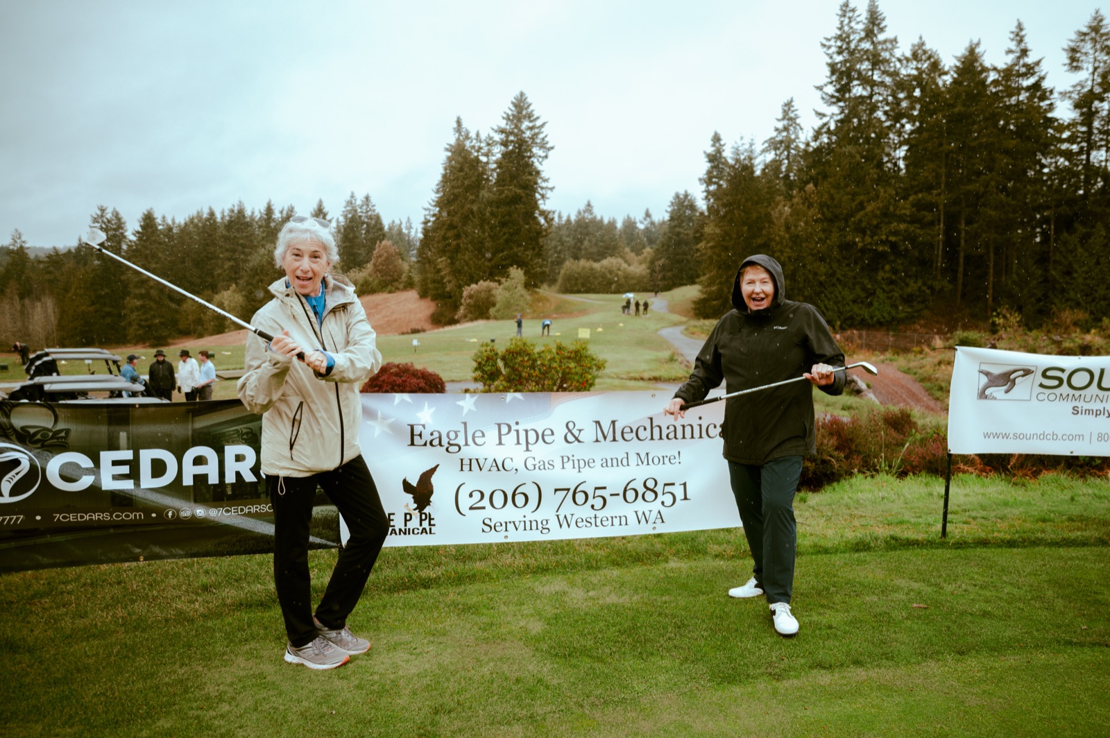 Two golfers laughing with clubs raised in the rain in front of the Eagle Pipe sponsor banner